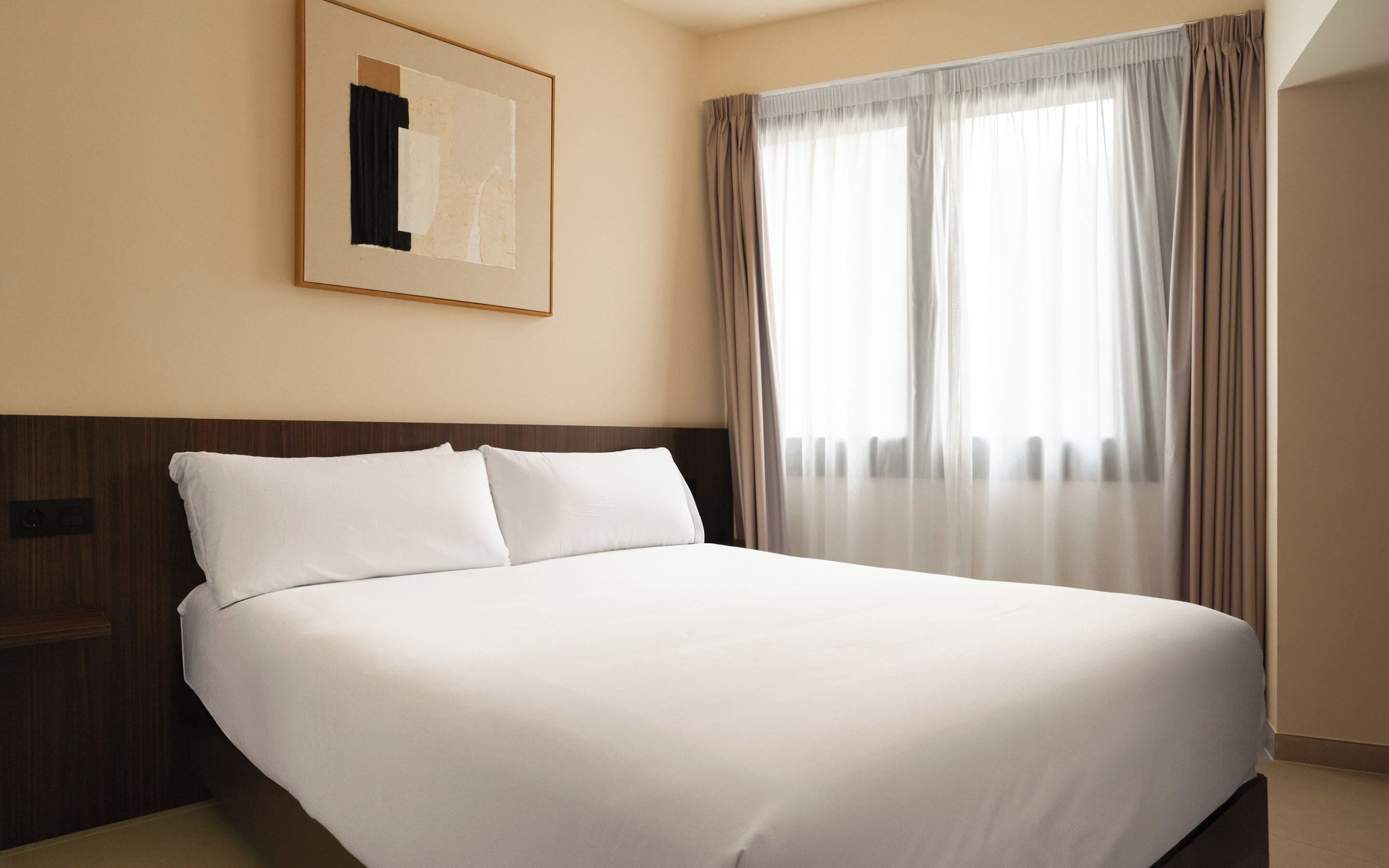 Una cama bien hecha en una habitación de hotel, con una obra enmarcada encima y cortinas translúcidas en la ventana.