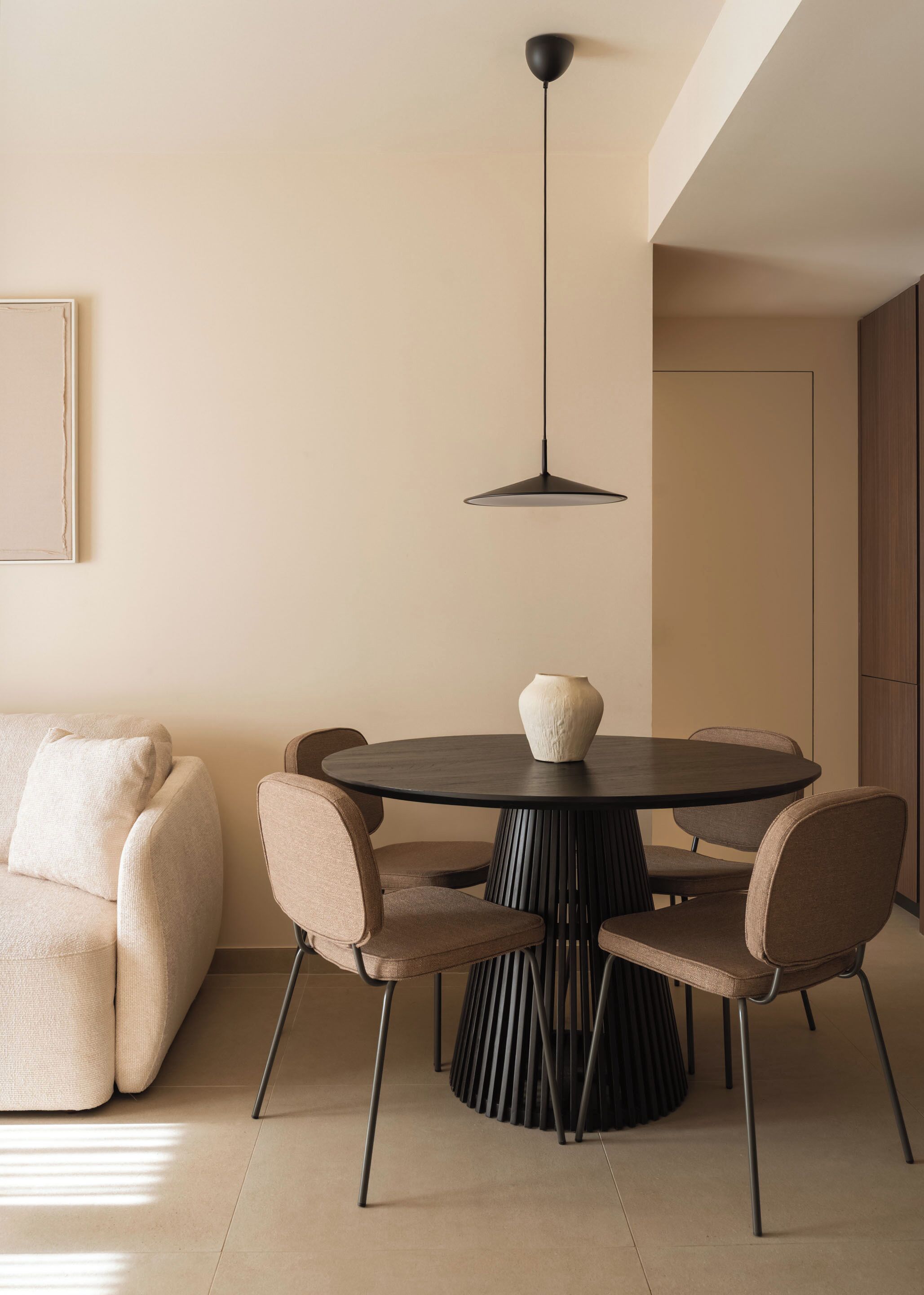 Un espace repas moderne avec une table ronde, six chaises, un vase posé sur la table, un luminaire suspendu et un canapé agrémenté de coussins.