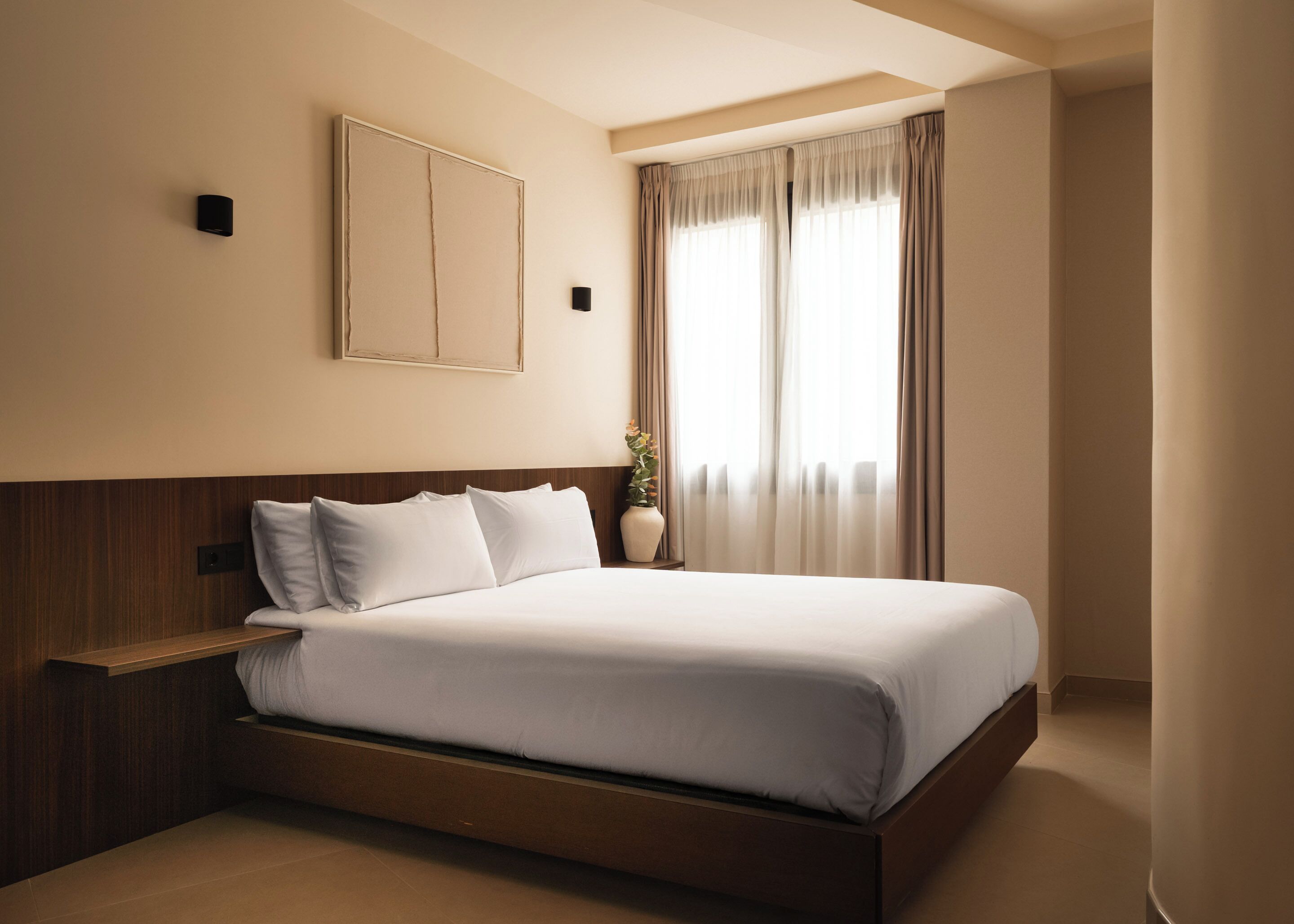 A hotel room with a bed, a bedside table, a wall-mounted light, and a window with curtains.