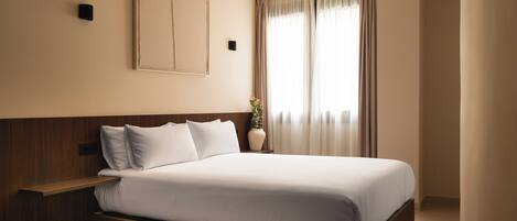 A hotel room with a bed, a bedside table, a wall-mounted light, and a window with curtains.