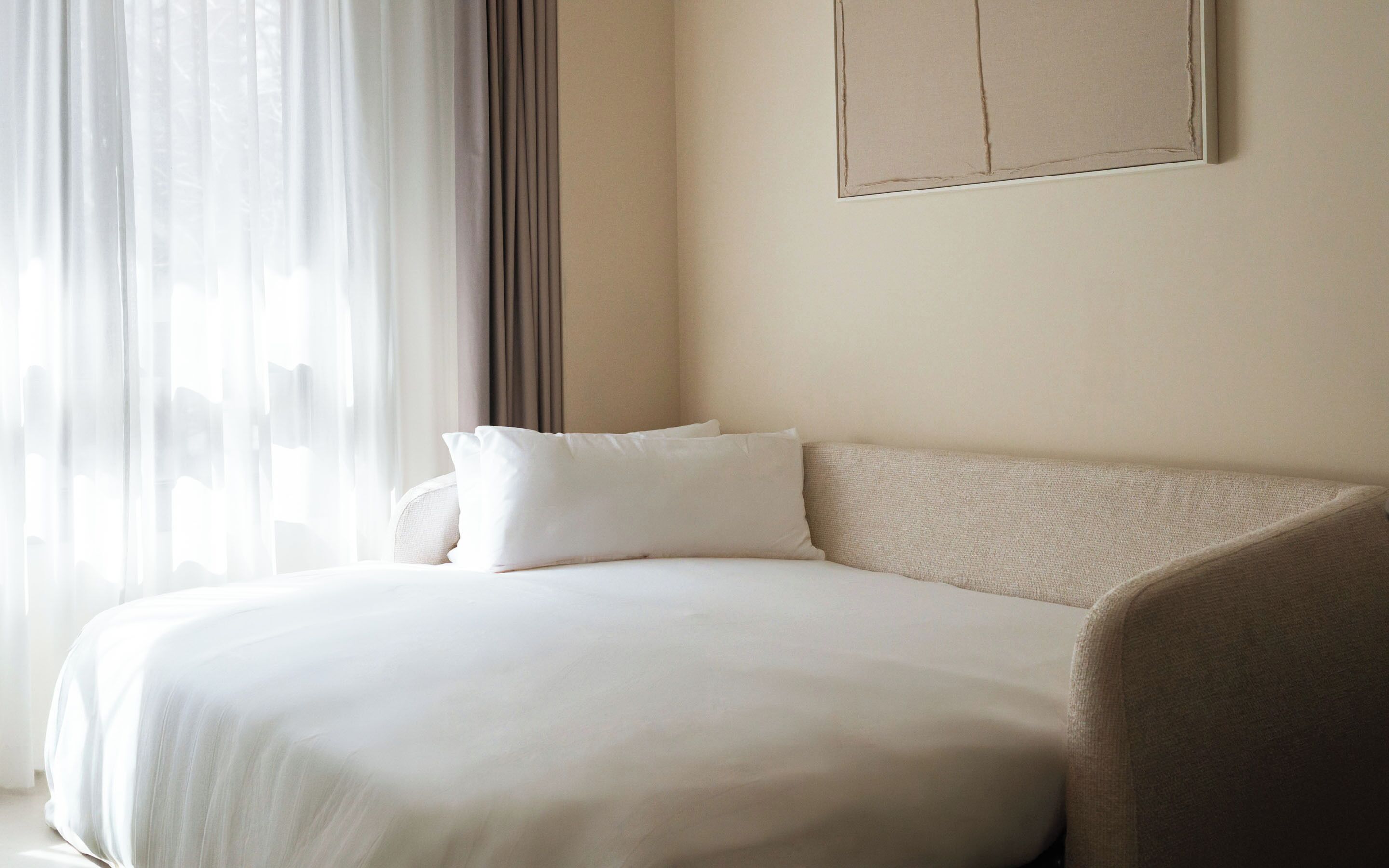 Una cama bien hecha con ropa blanca, un sofá color crema y un ventanal con cortinas translúcidas.