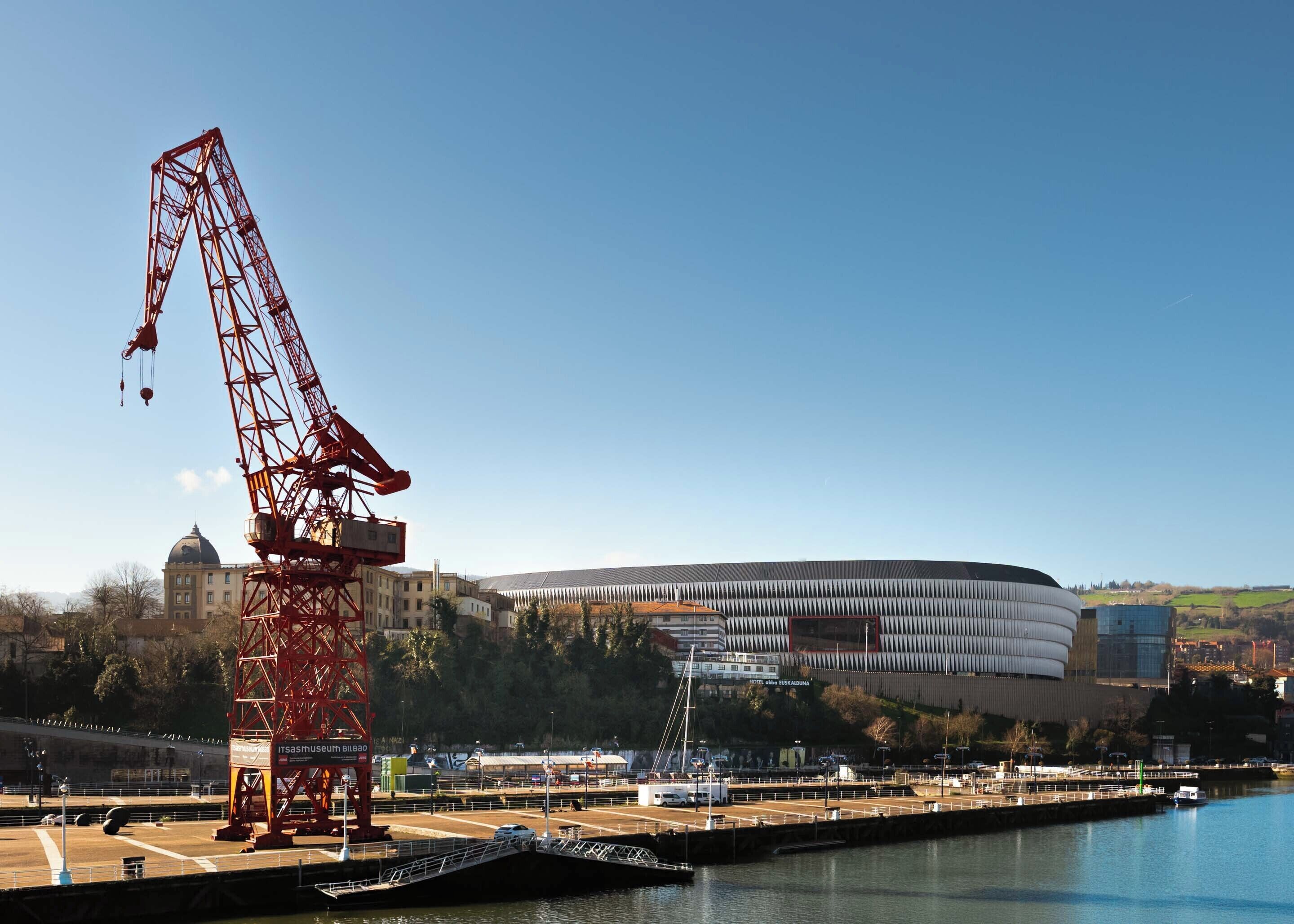 A large red crane, a modern building with a curved roof, and a body of water.