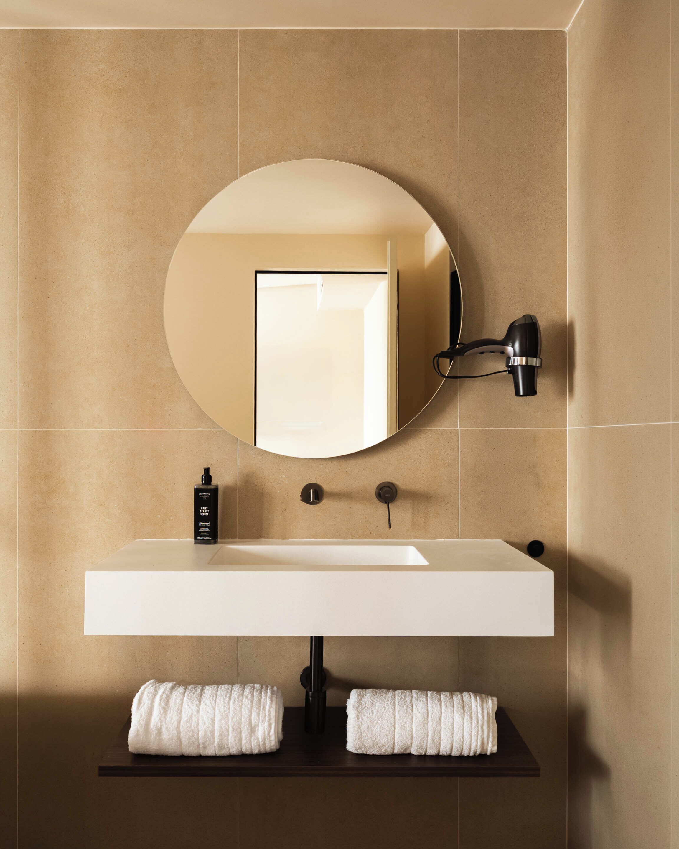 Une salle de bain avec un lavabo blanc, un miroir rond, un porte-serviettes et un sèche-cheveux.