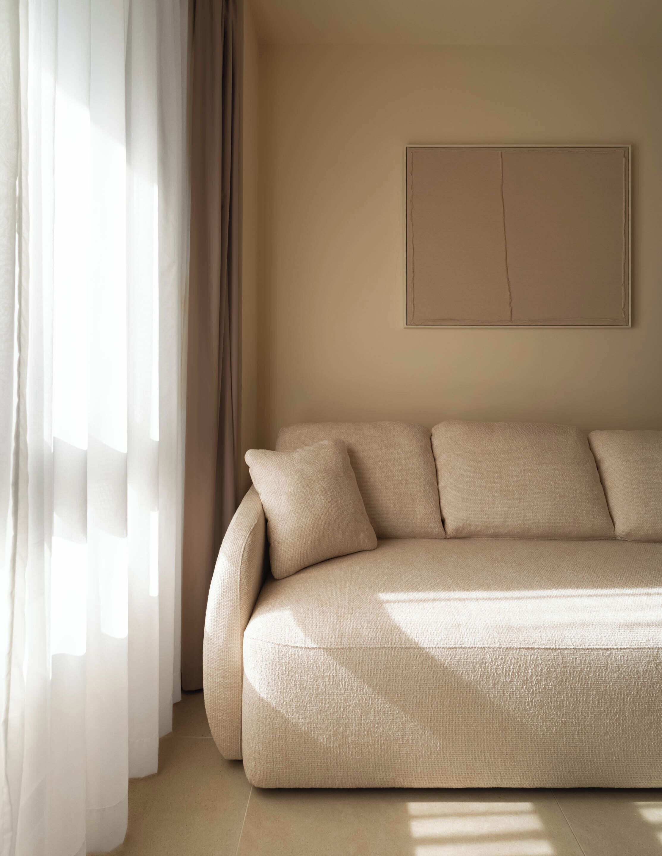 A minimalist living room with a beige sofa, sheer curtains, and a simple wall-mounted artwork.