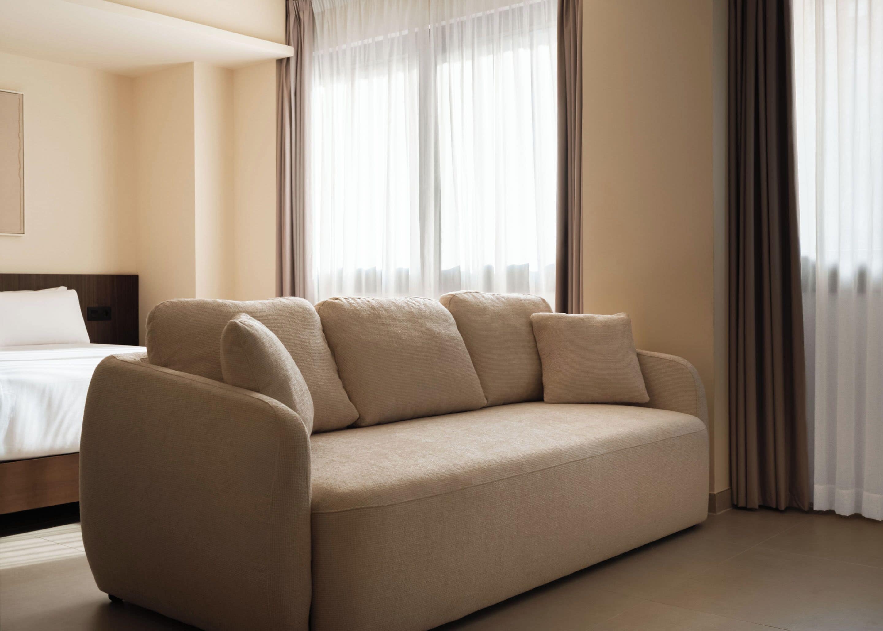 Une chambre d’hôtel moderne avec un canapé beige, un lit et une grande fenêtre avec des rideaux.