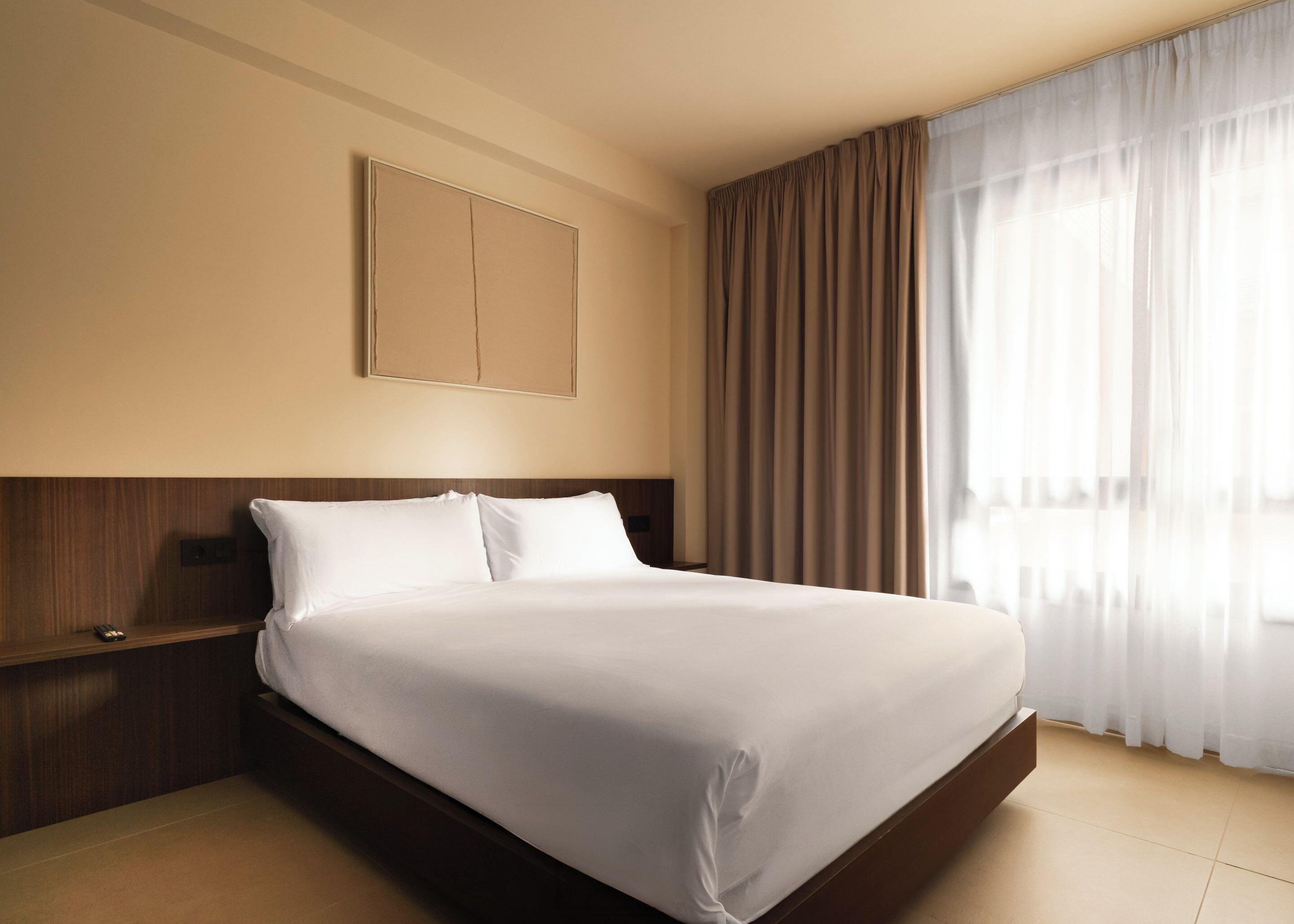 Una habitación de hotel con una cama grande, cabecera de madera, mesita de noche, televisor montado en la pared y una ventana con cortinas.