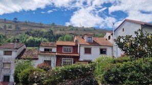 Uma vila no topo de uma colina com casas de telhas de barro tradicionais, muros de pedra e vegetação exuberante.