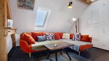 Ein gemütliches Zimmer mit Holzbalkendecke, einer Couch mit orangenen Kissen, einem kleinen Tisch und einem Fenster mit Blick.