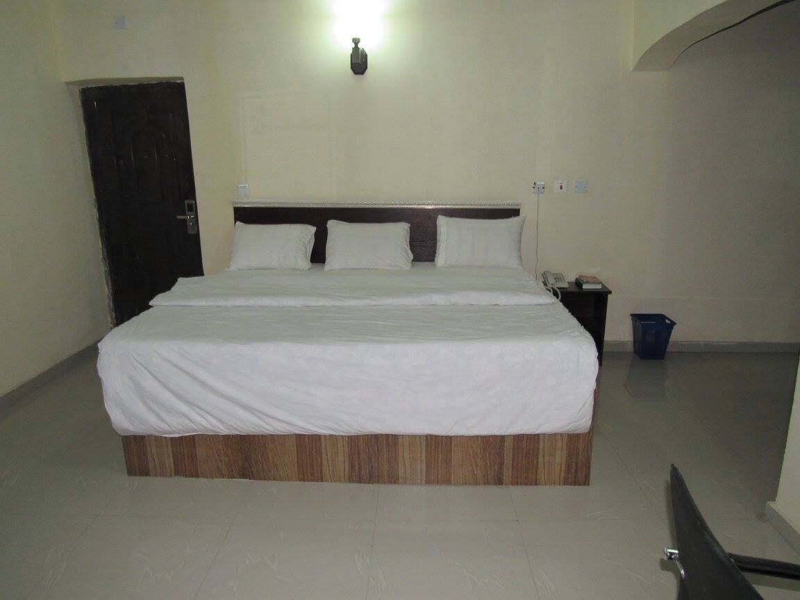 Quarto de hotel com uma cama grande, cabeceira de madeira, mesa de cabeceira com telefone e uma lixeira azul.