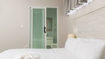 A hotel room with a bed, a bathroom visible through a glass door, and a headboard with curtains.