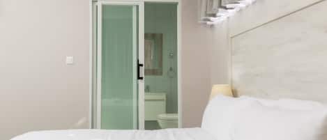 Habitación de hotel con cama, baño visible a través de una puerta de vidrio y cabecera con cortinas.