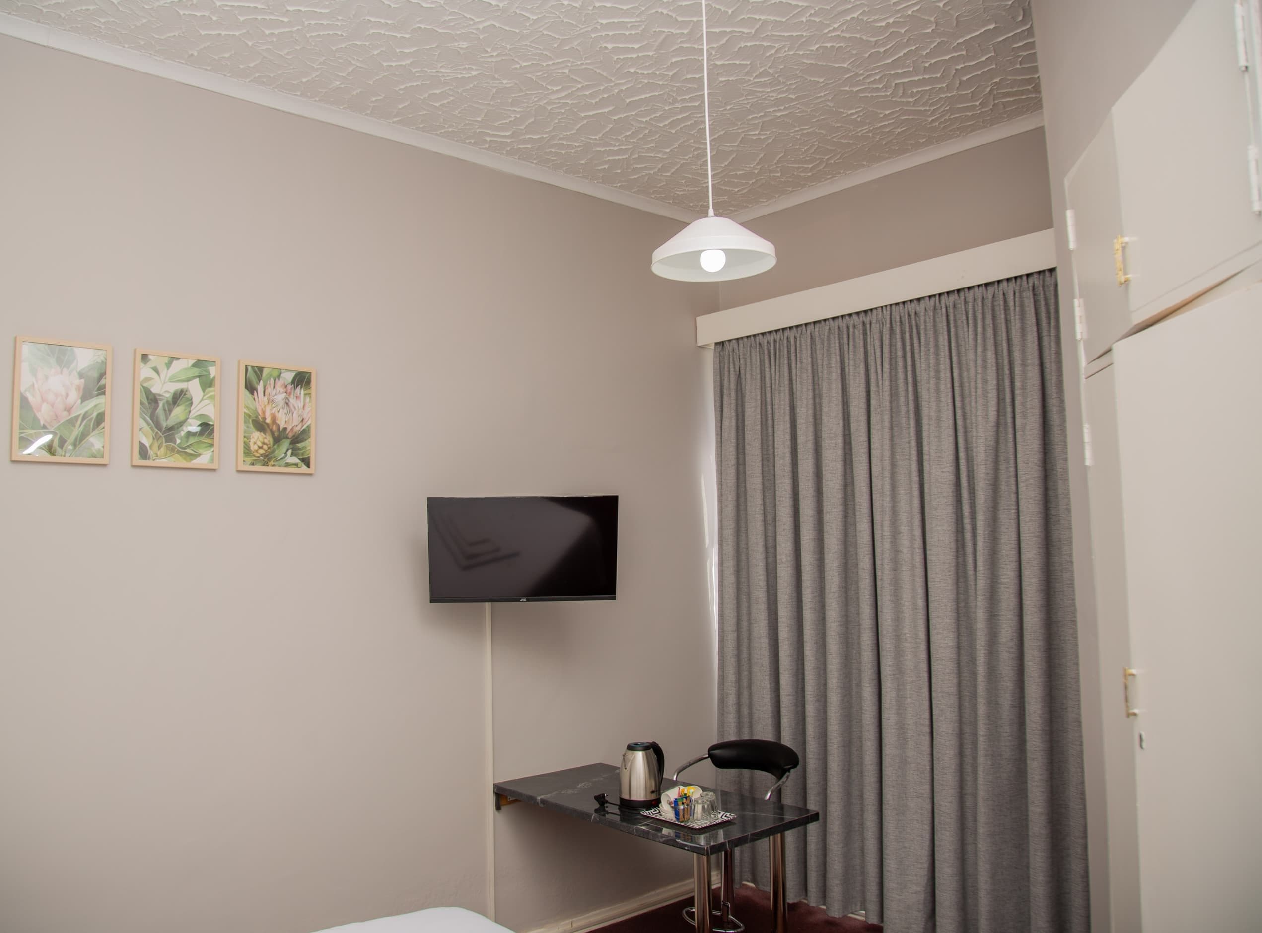 A room with a bed, a flat-screen TV mounted on the wall, a small table with a kettle, and a grey curtain.