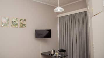 A room with a bed, a flat-screen TV mounted on the wall, a small table with a kettle, and a grey curtain.