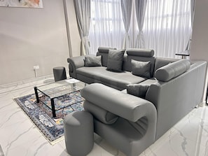 A modern living room with a grey sofa, ottoman, and a glass coffee table with a patterned rug. - Castle Rock Guest House  (Bulawayo)