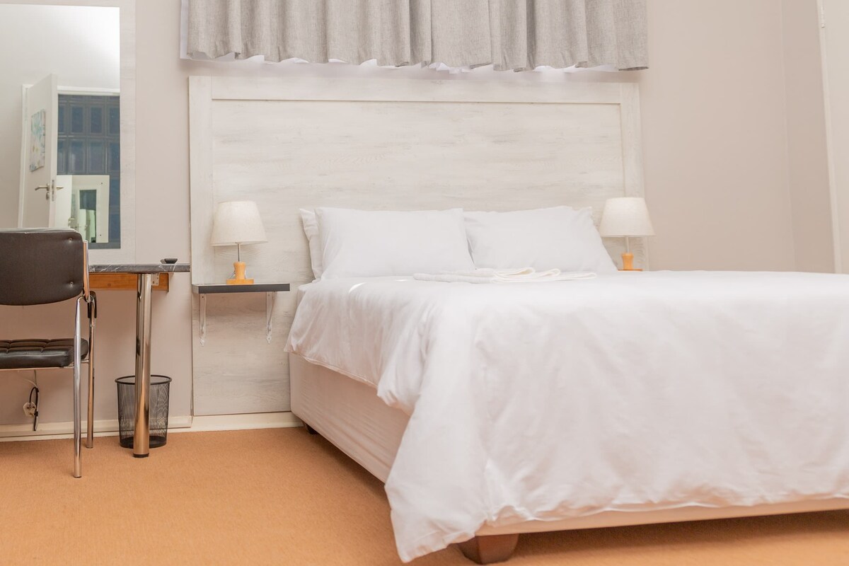A bedroom with a bed, two bedside tables with lamps, a chair, a small table, and a curtain.