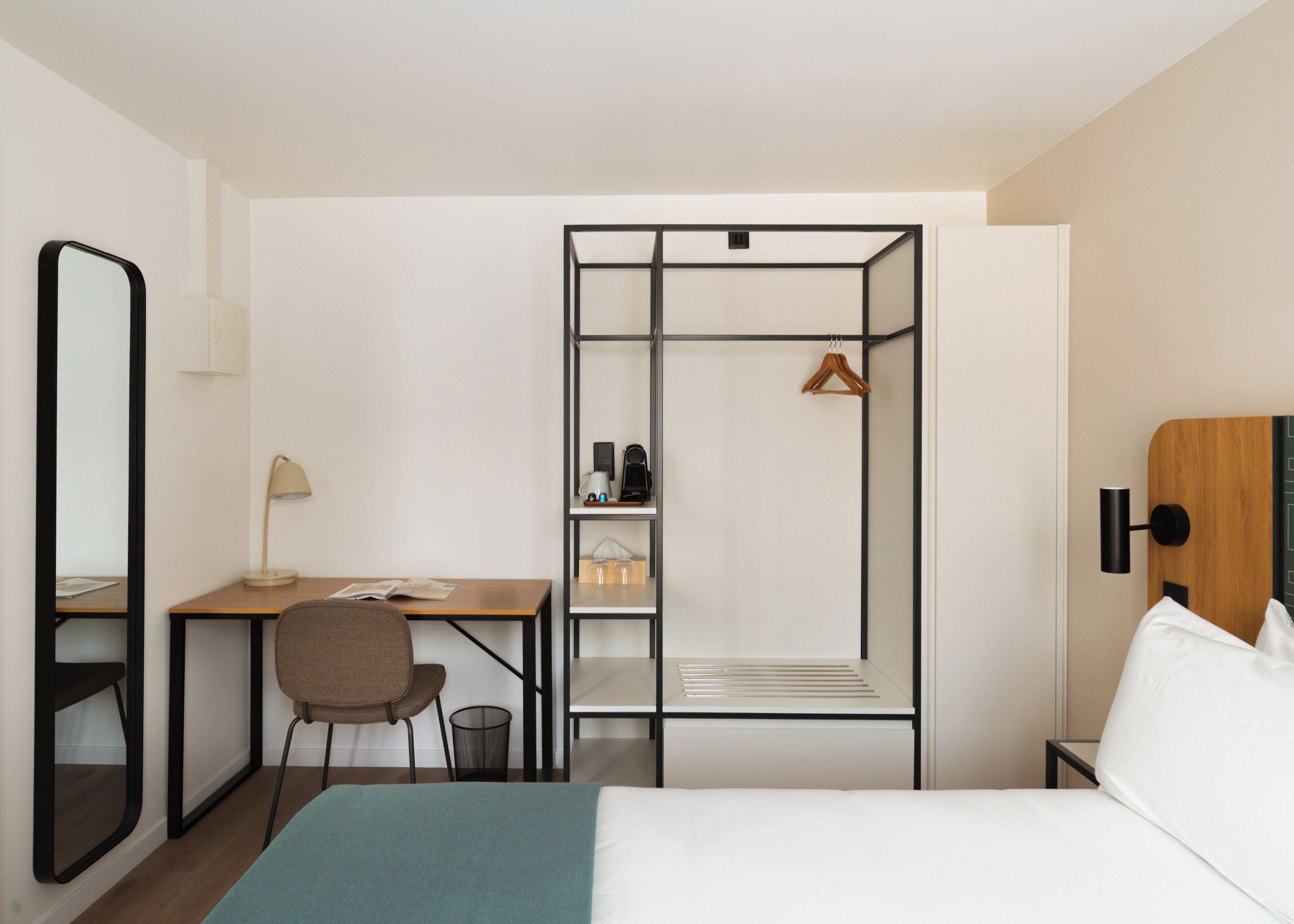 A hotel room with a bed, a desk with a chair, a wardrobe, a mirror, and a bedside table.