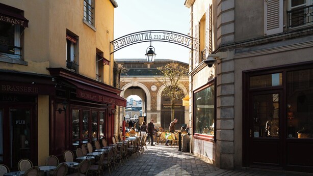 A narrow street with outdoor seating at cafes, a historic archway, and a mix of commercial and residential buildings. - Limehome Versailles Rue du Bailliage (Versailles)