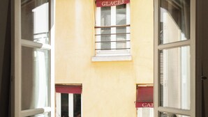 An open window with a view of a building with a 'GLACES' sign, a metal railing, and a red awning. - Limehome Versailles Rue du Bailliage (Versailles)