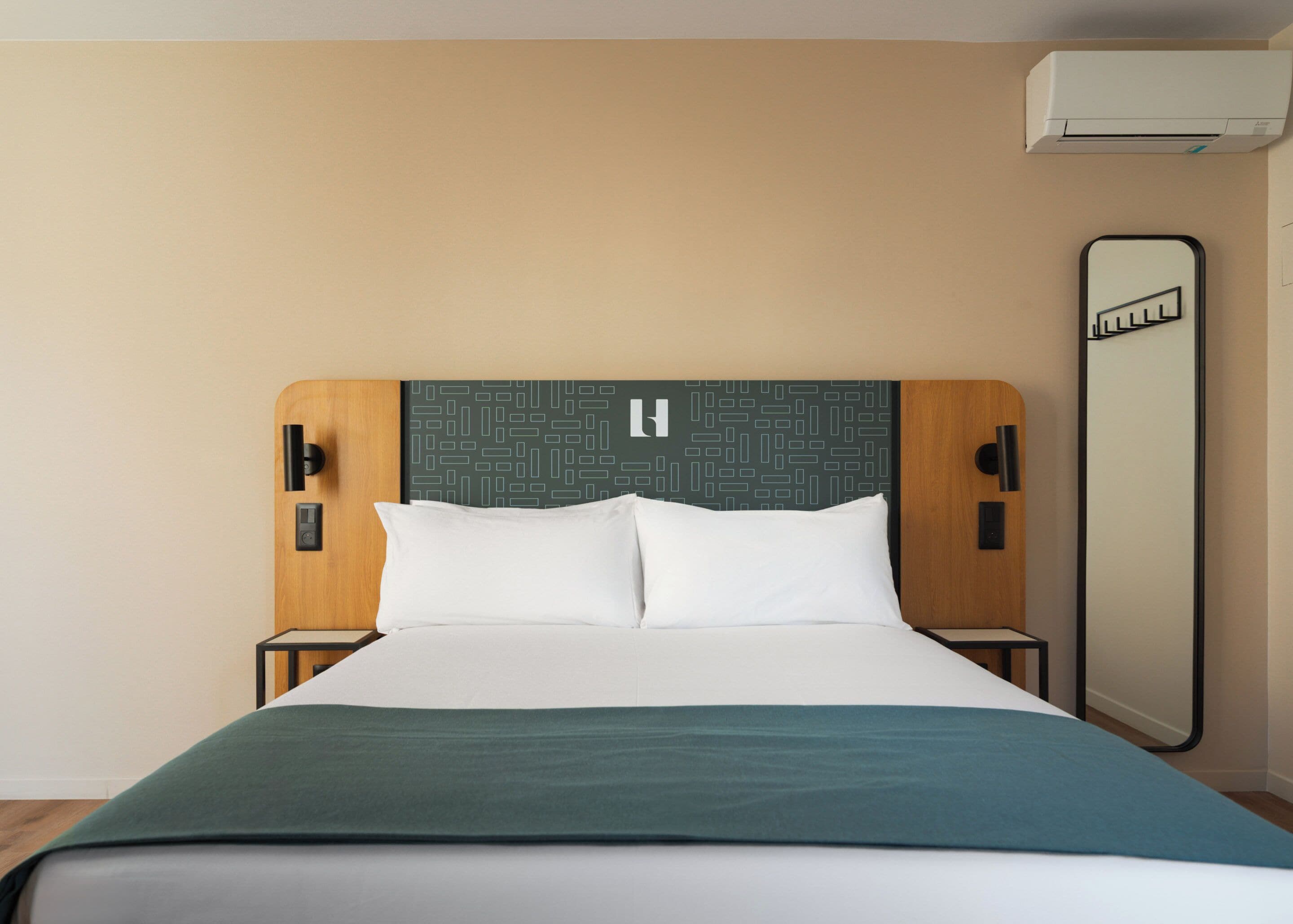A hotel room with a bed, a headboard with a geometric pattern, a bedside table, a nightstand, and a large mirror.
