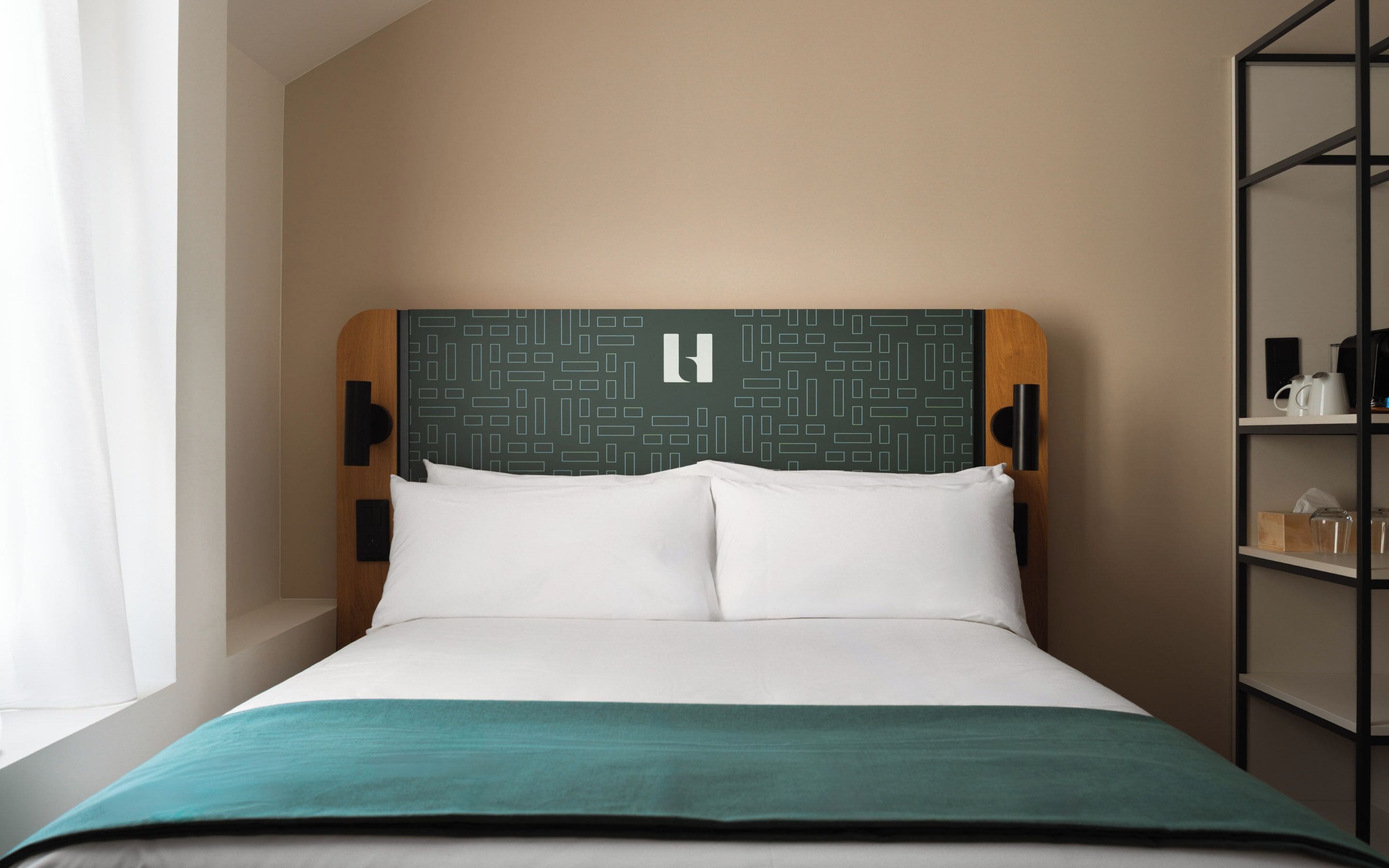 Habitación de hotel con cama, cabecero de estampado geométrico y una manta azul verdosa. Se ve una estantería con objetos a la derecha.