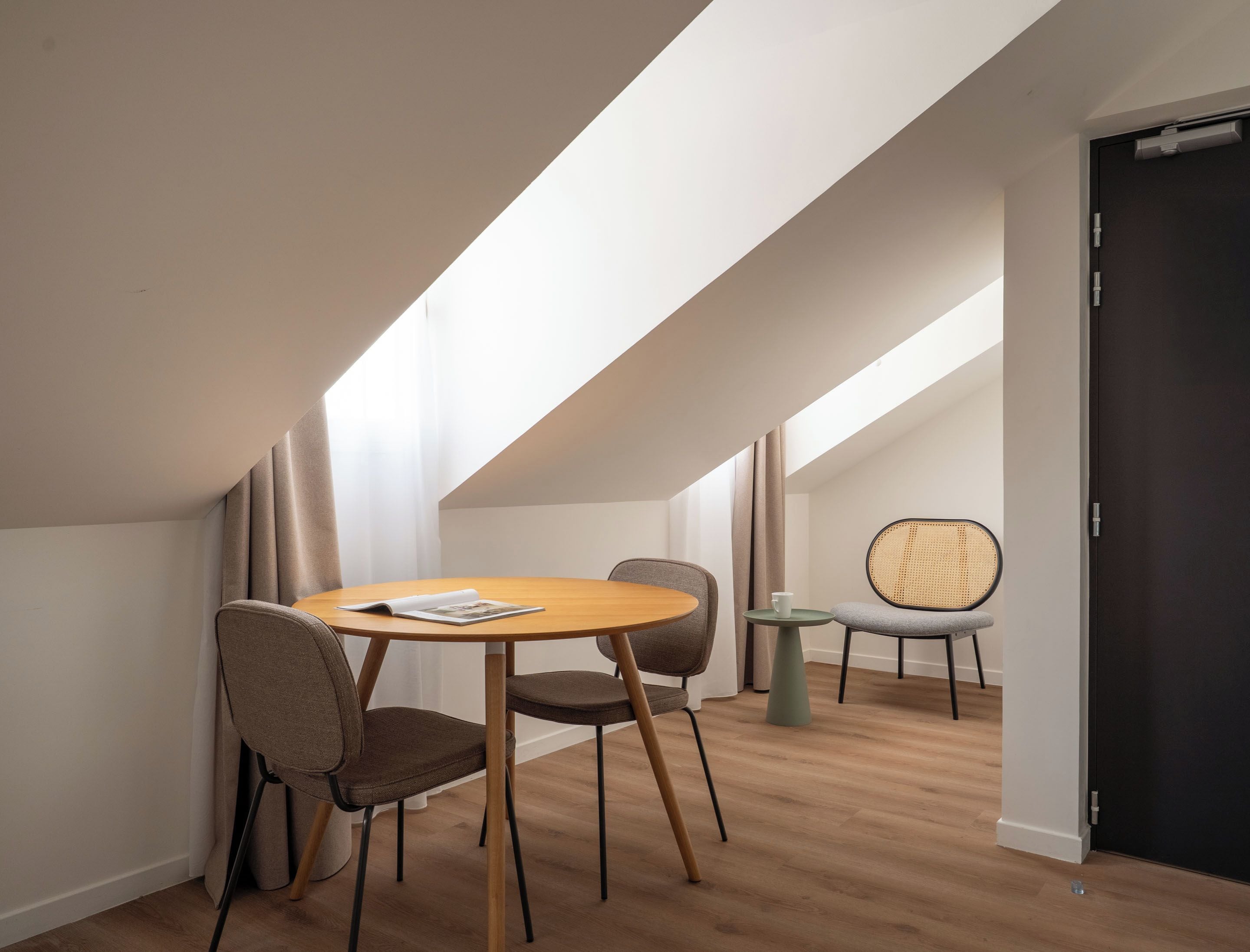 A modern loft bedroom with a sloped ceiling, a round wooden dining table, two chairs, a small side table, and a rattan chair.