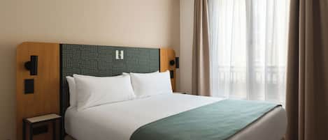 Habitación de hotel con cama, mesita de noche, panel de control en la pared y ventana con cortinas.
