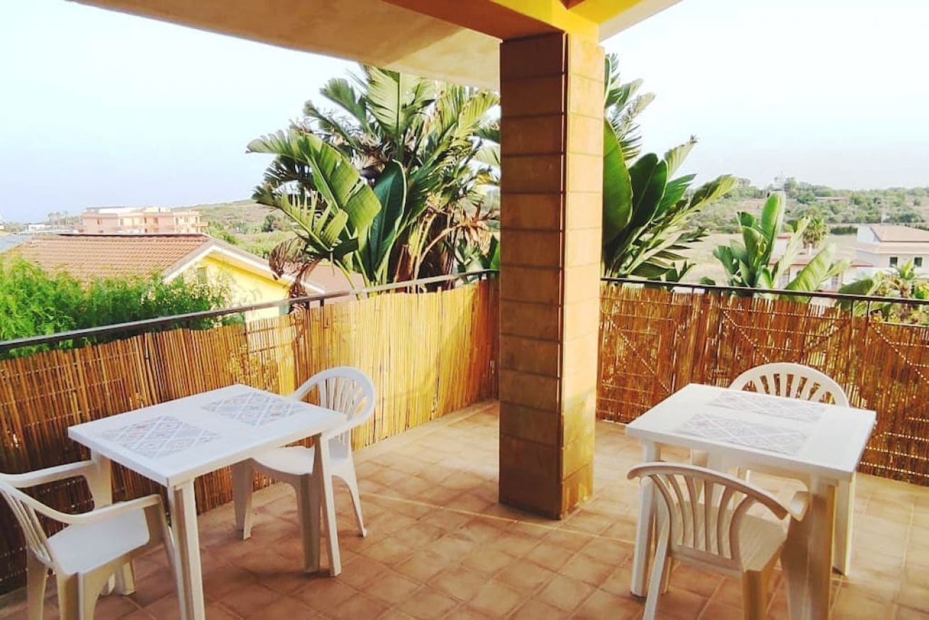 Een balkon met twee witte tafels en stoelen, een bamboe hek en uitzicht op gebouwen en groen.