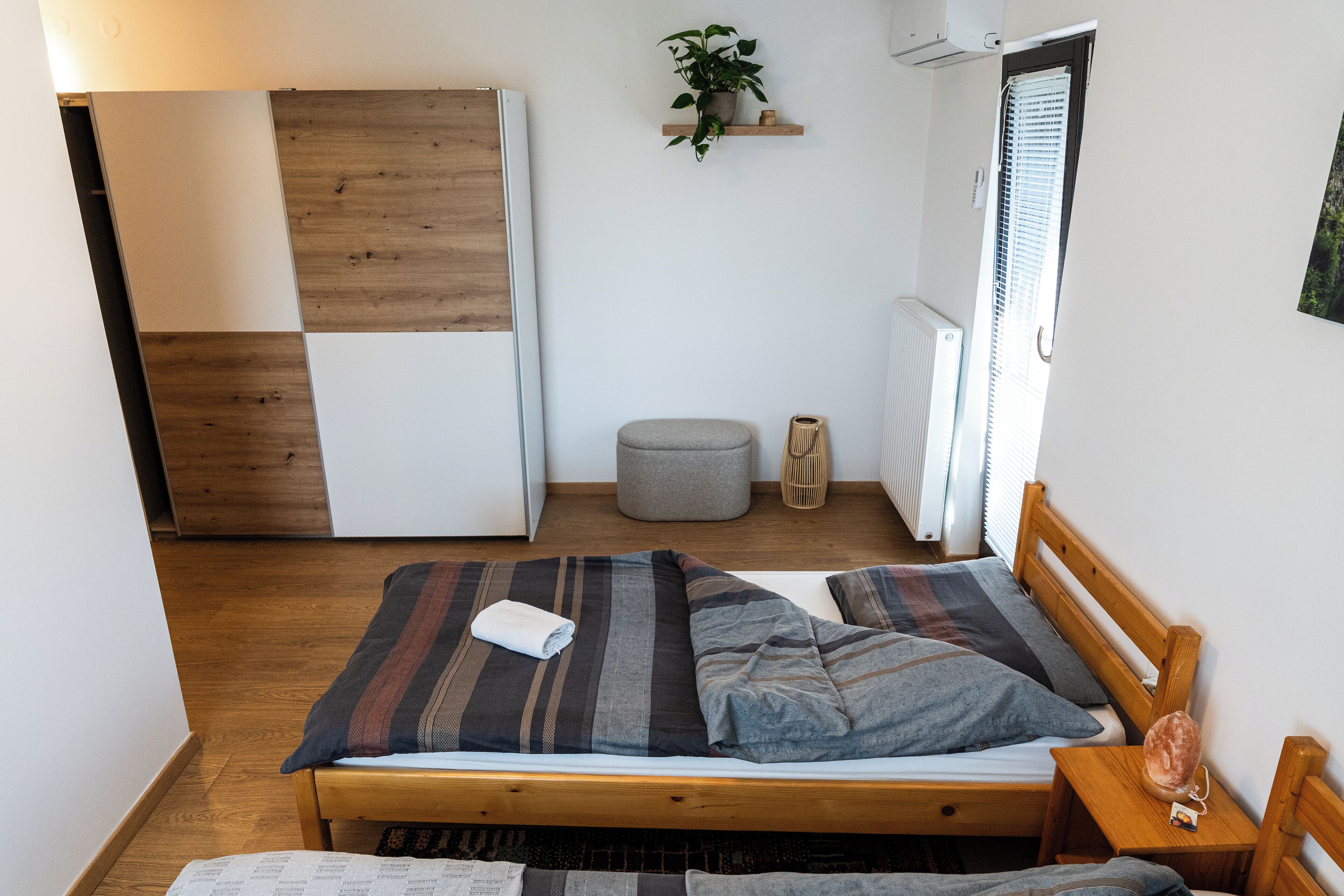 A compact bedroom with a wooden bed, a small bedside table, a grey ottoman, a wall-mounted shelf with a plant, and a radiator.