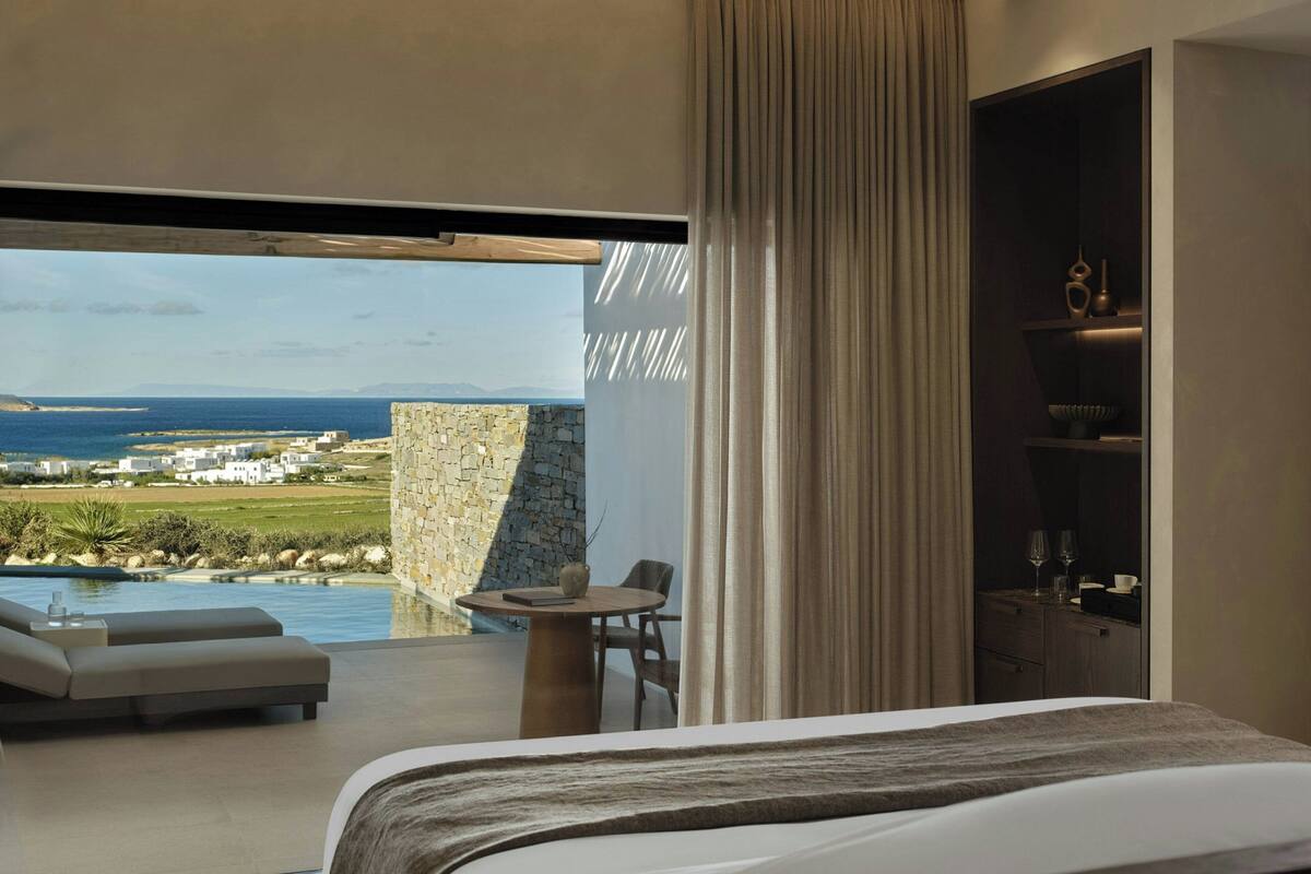 A modern hotel room with a large bed, a balcony with a view of the sea, and a dining area with a table and chairs.