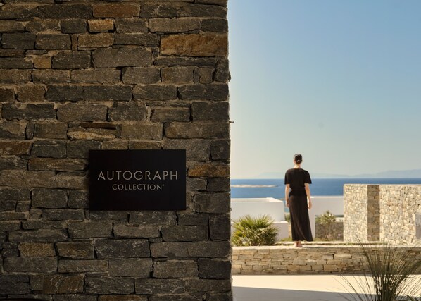 A stone wall with a sign reading 'AUTOGRAPH COLLECTION' and a person standing by a building with a view of the sea. - Oroséa, Paros, Autograph Collection - Adults Only (Paros)