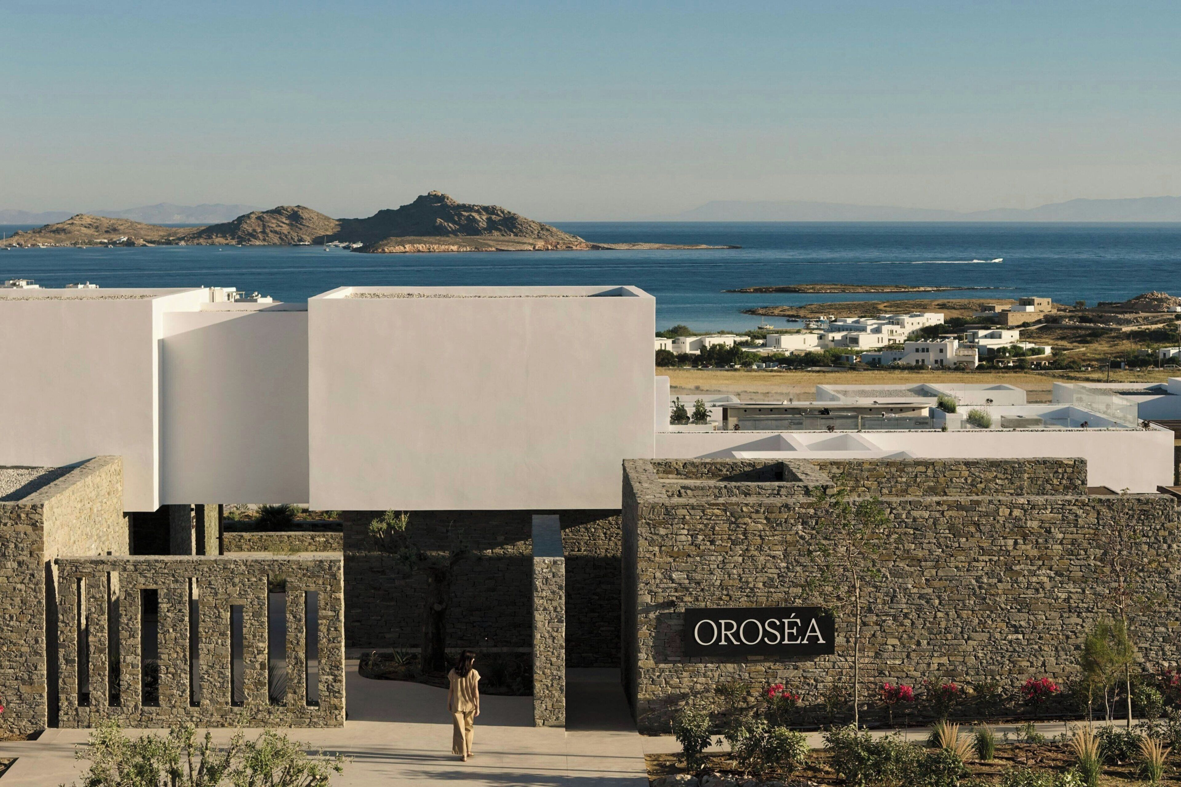 A coastal property with a stone wall entrance, a sign reading 'Oroséa,' and a view of the sea and distant hills.