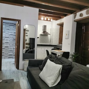 Un salon moderne avec un canapé foncé, un miroir et un coin cuisine doté de carreaux blancs et d’un plan de travail noir.