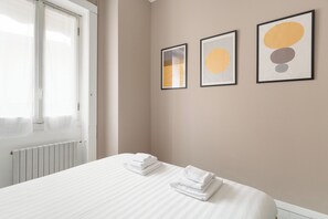 A neatly made bed with folded white towels, a window with sheer curtains, and three framed abstract art pieces on the wall. - Brera Apartments in Sant'Ambrogio (Milan)