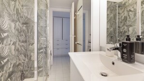 Un baño con un lavabo blanco, un espejo y una ducha con puerta de cristal. Hay dos botellas de champú sobre el lavabo.