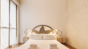 Una cama bien hecha con toallas dobladas, dos lámparas de noche y un ventanal con cortinas.