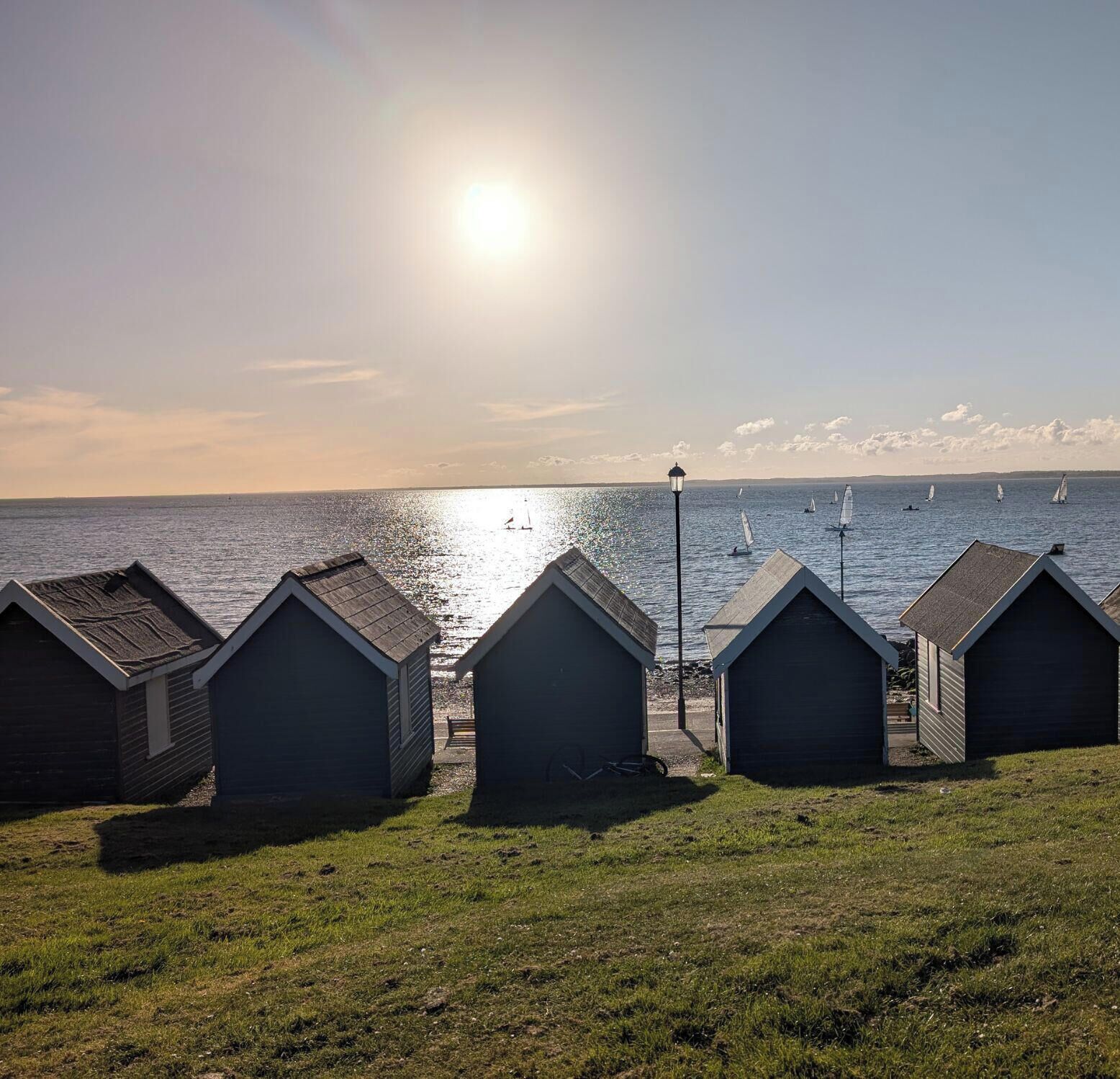 A row of beach huts with a view of the sea and sailboats.