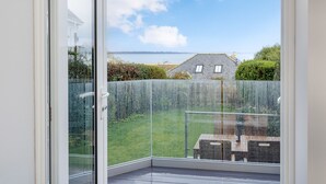 Ein Balkon mit Glastür, Blick auf eine grüne Fläche und ein entferntes Gebäude über dem Wasser.