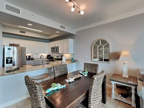 A modern kitchen with white cabinets, stainless steel appliances, and a wooden dining table with woven chairs. - The Indies #209 | Quiet Beach, Pool, Hot Tub, Gym, Rooftop Deck (Gulf Shores)