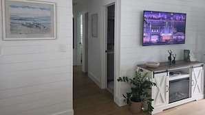 A hallway with a framed painting, a television displaying a cityscape, and a potted plant. - Charming Beach Cottage with Hot Tub only 1 Mile to Gulf! (Naples)