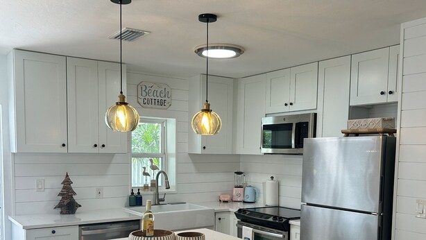 A modern kitchen with white cabinetry, a central island with bar stools, pendant lighting, and a stainless steel refrigerator. - Charming Beach Cottage with Hot Tub only 1 Mile to Gulf! (Naples)