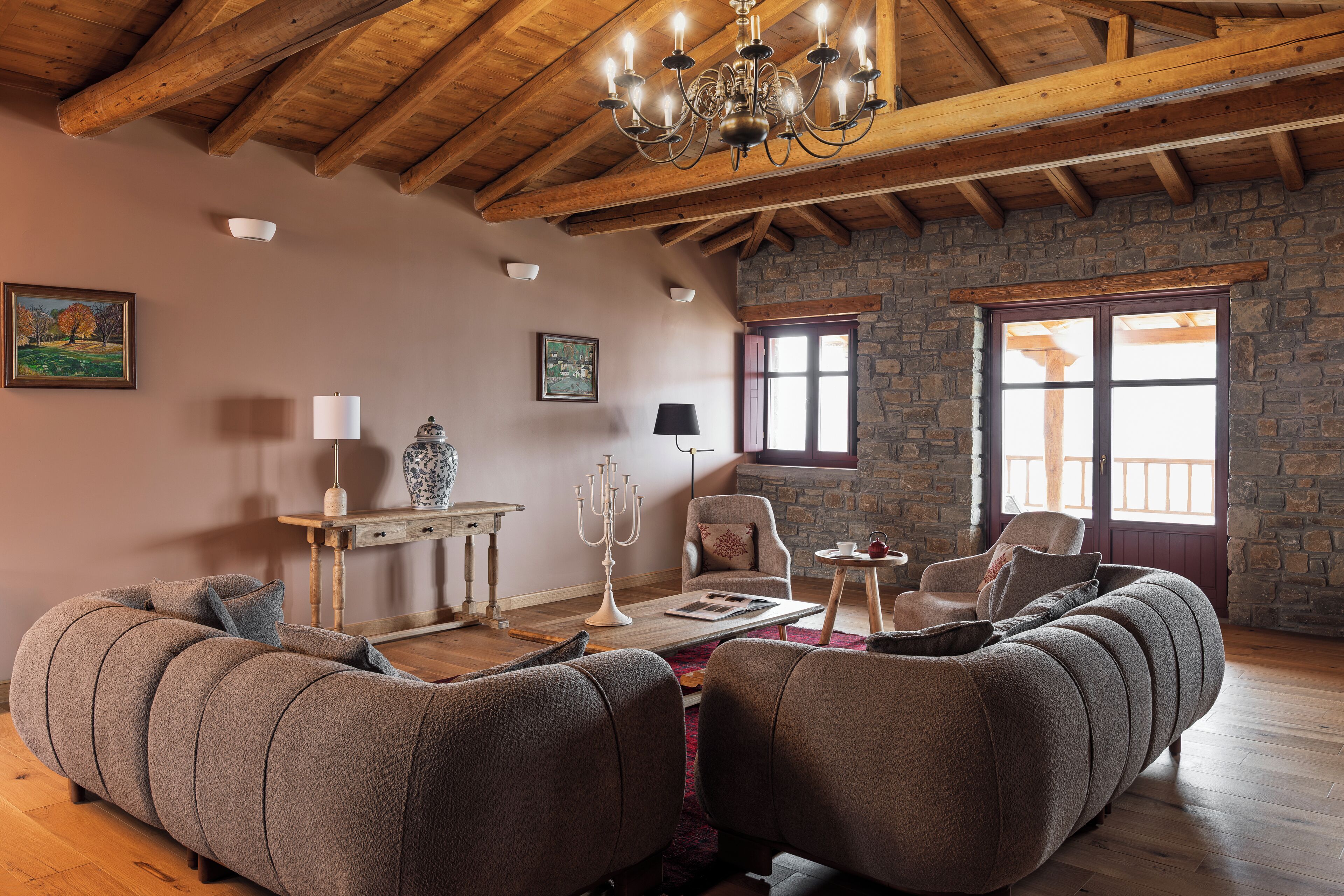 Un salon chaleureux avec un mur en pierre, des poutres apparentes en bois au plafond, un lustre, un grand canapé, une table basse et une cheminée.