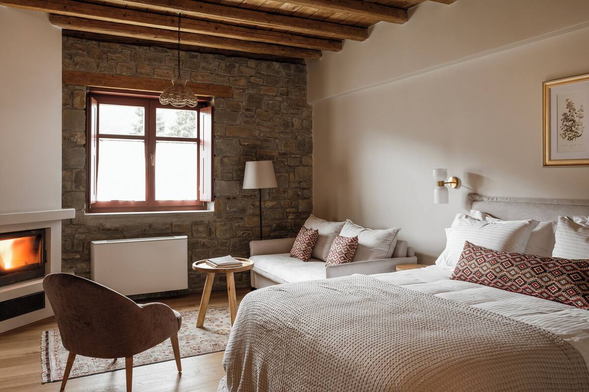 A cozy bedroom with a stone wall, a fireplace, a bed with patterned pillows, a side table, and a chair.