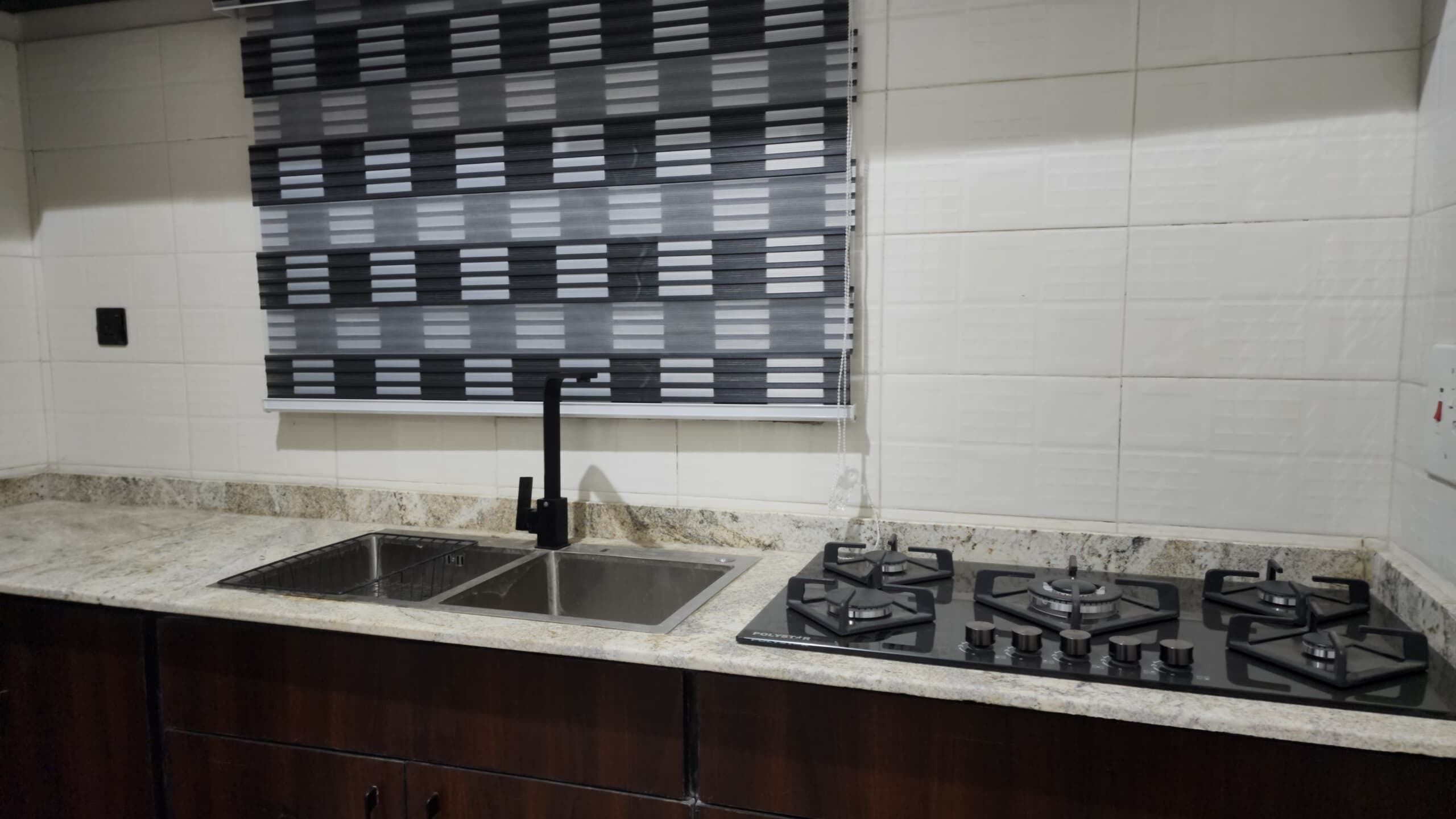A kitchen with a double sink, a gas stove, and a black and white patterned window blind.