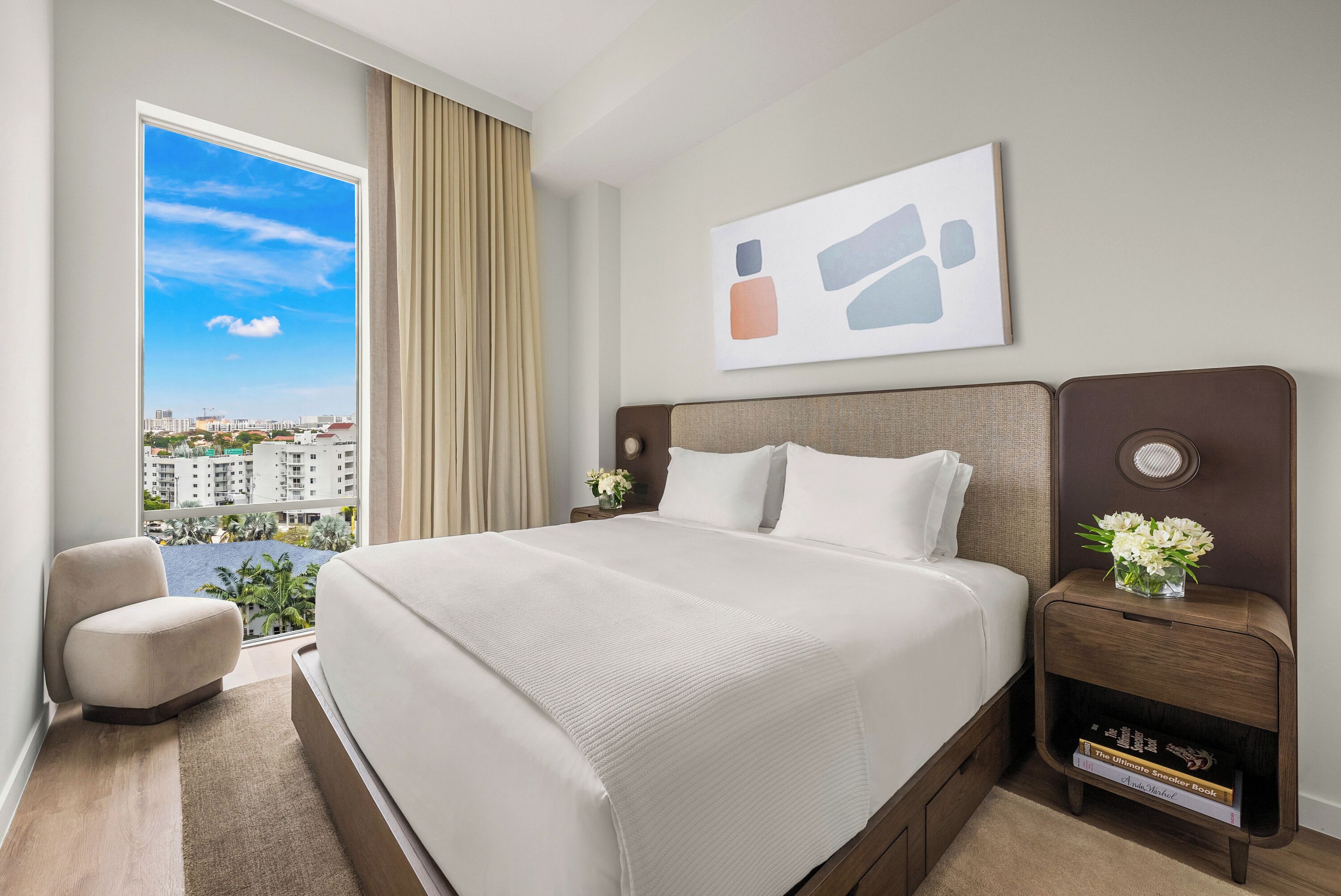 A hotel room with a large bed, a nightstand with a vase of flowers, a view of buildings through a window, and a painting on the wall.