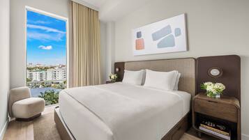 A hotel room with a large bed, a nightstand with a vase of flowers, a view of buildings through a window, and a painting on the wall.