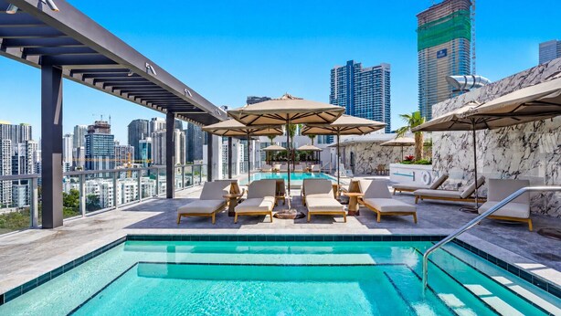 Un área de piscina en la azotea con sillones de descanso y sombrillas, con vistas al horizonte de la ciudad.