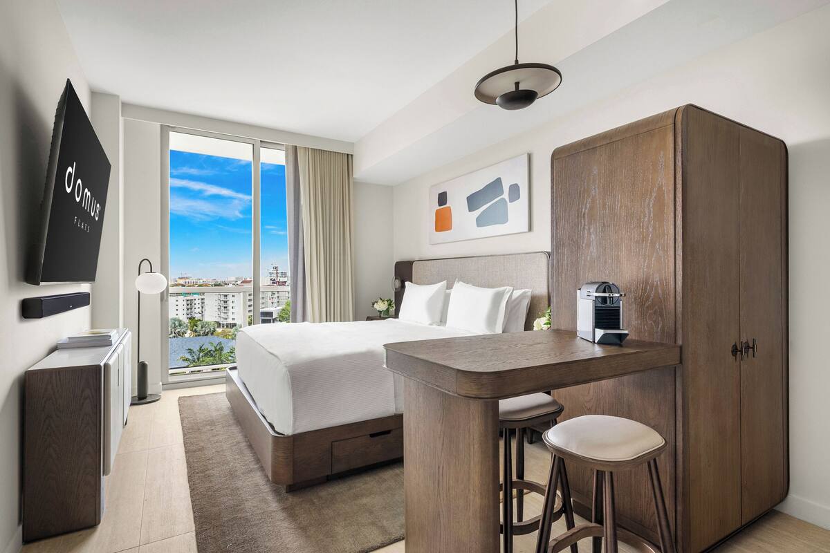 Habitación de hotel con cama, escritorio con dos taburetes, armario, televisor y ventana grande con vista a la ciudad.