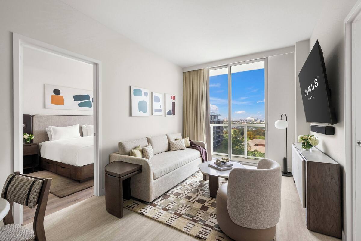 Una habitación de hotel moderna con cama, sofá, sillón y un amplio ventanal con vistas a la ciudad.