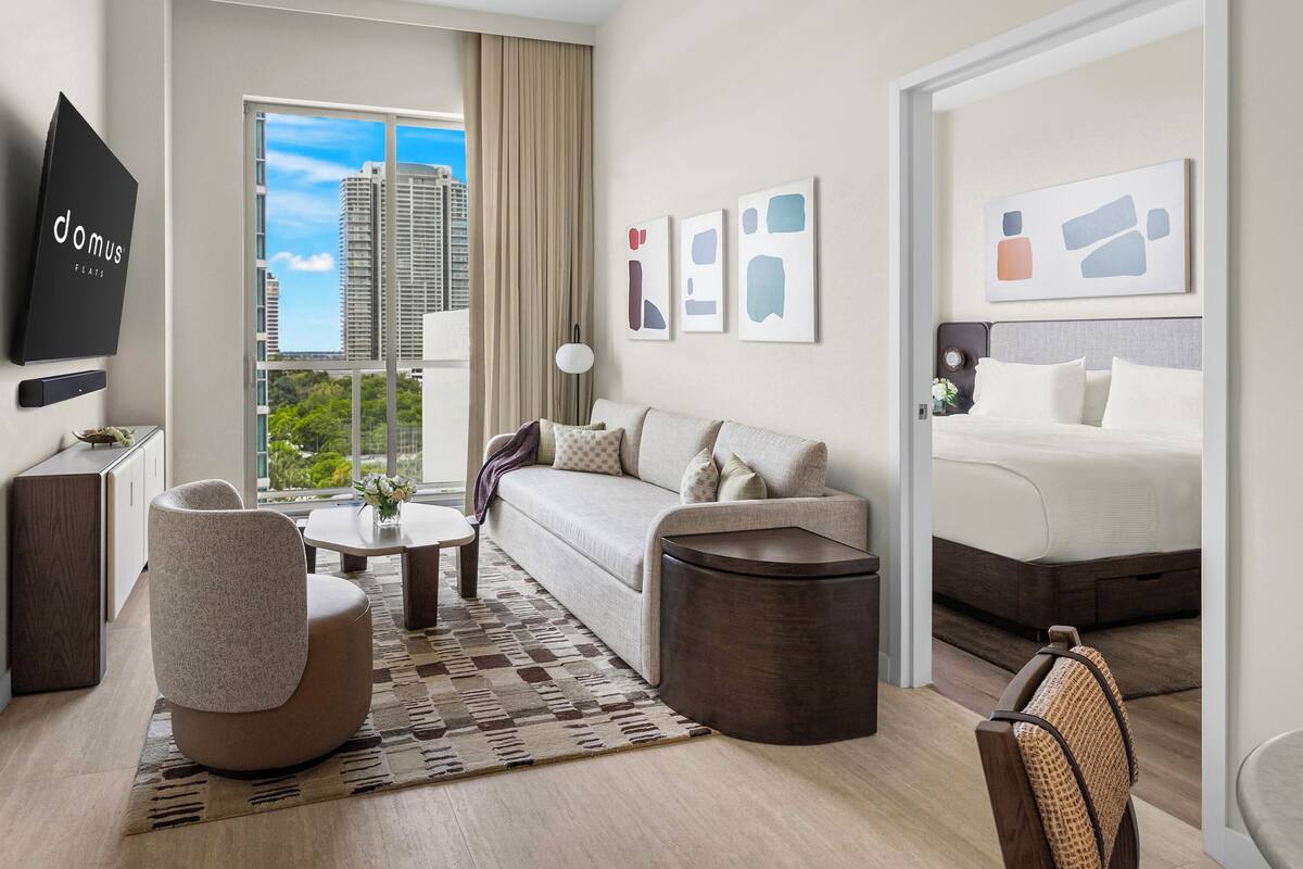 Una habitación de hotel moderna con sofá, una mesa redonda, una silla, cama y televisor en la pared.