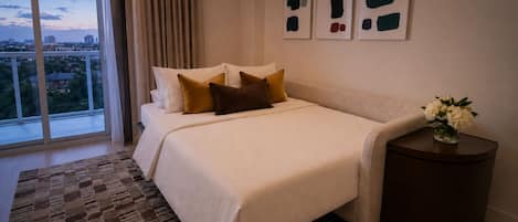A hotel room with a large window, a bed with multiple pillows, a small side table with a vase, and abstract wall art.